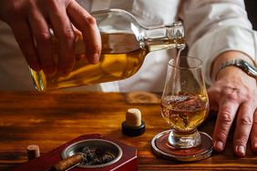 A person pours whiskey into a glass on a wooden table, next to a cigar 