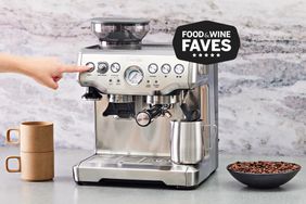 Hand pressing a button on the Breville Barista Express displayed with cups and a bowl with coffee on a grey counter